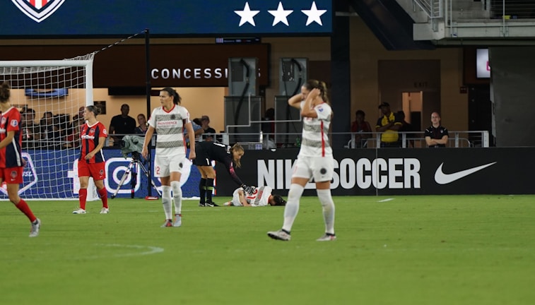 A soccer match scene shows players on a field, some in red and others in white uniforms. One player is injured and lying on the ground while another kneels beside them. The area behind features a concession stand with a large advertisement for Nike Soccer. Spectators and staff are visible in the background.