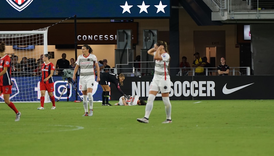 A soccer match scene shows players on a field, some in red and others in white uniforms. One player is injured and lying on the ground while another kneels beside them. The area behind features a concession stand with a large advertisement for Nike Soccer. Spectators and staff are visible in the background.