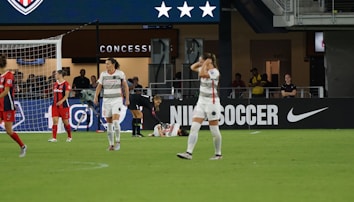 A soccer match scene shows players on a field, some in red and others in white uniforms. One player is injured and lying on the ground while another kneels beside them. The area behind features a concession stand with a large advertisement for Nike Soccer. Spectators and staff are visible in the background.