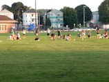 A group of people engaged in various physical activities on a grassy field surrounded by a few residential buildings and trees. Some individuals are stretching, while others are either sitting or standing. There is a blue playground slide visible in the background, and the setting appears to be in a park or sports field during daylight.