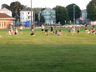 A group of people engaged in various physical activities on a grassy field surrounded by a few residential buildings and trees. Some individuals are stretching, while others are either sitting or standing. There is a blue playground slide visible in the background, and the setting appears to be in a park or sports field during daylight.