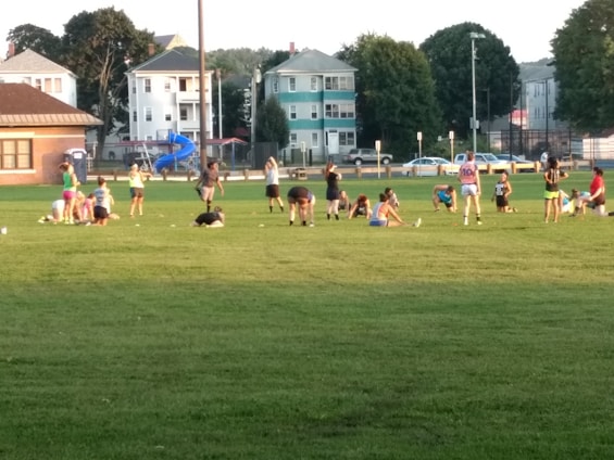 A group of people engaged in various physical activities on a grassy field surrounded by a few residential buildings and trees. Some individuals are stretching, while others are either sitting or standing. There is a blue playground slide visible in the background, and the setting appears to be in a park or sports field during daylight.
