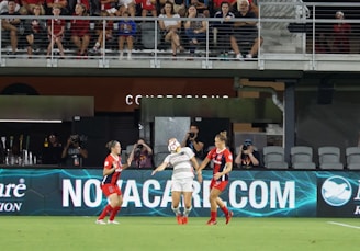 A group of female soccer players are actively engaged in a match on the field. One player is in mid-action, heading the ball, while two others are nearby, watching attentively. The audience is seated in the stands in the background, some cheering or capturing the moment with cameras.