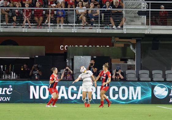 A group of female soccer players are actively engaged in a match on the field. One player is in mid-action, heading the ball, while two others are nearby, watching attentively. The audience is seated in the stands in the background, some cheering or capturing the moment with cameras.
