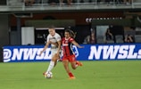 Two female soccer players are in action on a soccer field. One is wearing a white jersey with white shorts, and the other is in a red and black striped jersey with red shorts. They appear focused as they vie for control of the soccer ball. The background shows a digital advertisement, audiences in the stands, and part of the stadium.