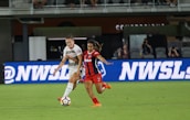 Two female soccer players are in action on a soccer field. One is wearing a white jersey with white shorts, and the other is in a red and black striped jersey with red shorts. They appear focused as they vie for control of the soccer ball. The background shows a digital advertisement, audiences in the stands, and part of the stadium.