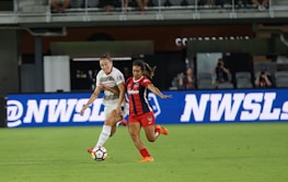 Two female soccer players are in action on a soccer field. One is wearing a white jersey with white shorts, and the other is in a red and black striped jersey with red shorts. They appear focused as they vie for control of the soccer ball. The background shows a digital advertisement, audiences in the stands, and part of the stadium.