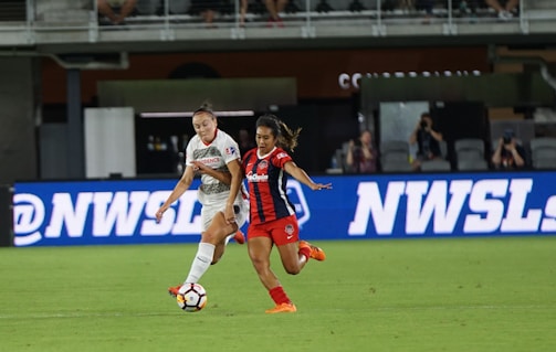 Two female soccer players are in action on a soccer field. One is wearing a white jersey with white shorts, and the other is in a red and black striped jersey with red shorts. They appear focused as they vie for control of the soccer ball. The background shows a digital advertisement, audiences in the stands, and part of the stadium.