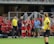A soccer team is gathered near the sidelines with players and coaches interacting. A player in a red and black uniform is clapping, and another wearing number 16 is high-fiving. A referee in a yellow shirt is standing nearby with substitution equipment. The benches are filled with people and the background shows stadium seating.