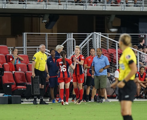 A soccer team is gathered near the sidelines with players and coaches interacting. A player in a red and black uniform is clapping, and another wearing number 16 is high-fiving. A referee in a yellow shirt is standing nearby with substitution equipment. The benches are filled with people and the background shows stadium seating.