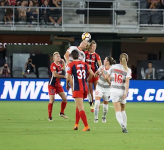 A group of female soccer players are on the field in the middle of an intense moment during a match. Two players are leaping to head the ball, with expressions of focus and determination. The scene is set in a stadium with spectators visible in the background.
