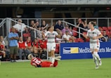 A soccer match is taking place with one player in a white and gray uniform raising her arms, possibly in celebration or frustration. Another player in red is on the ground. A third player in a white uniform runs across the field. Spectators are seated in the background watching the action.