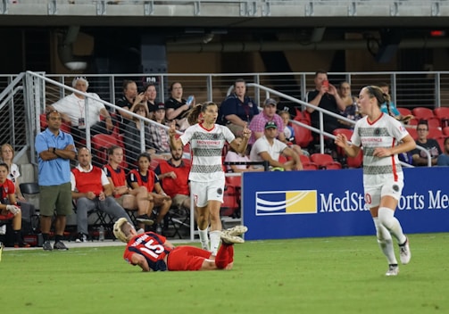 A soccer match is taking place with one player in a white and gray uniform raising her arms, possibly in celebration or frustration. Another player in red is on the ground. A third player in a white uniform runs across the field. Spectators are seated in the background watching the action.