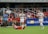 A soccer match is taking place with one player in a white and gray uniform raising her arms, possibly in celebration or frustration. Another player in red is on the ground. A third player in a white uniform runs across the field. Spectators are seated in the background watching the action.