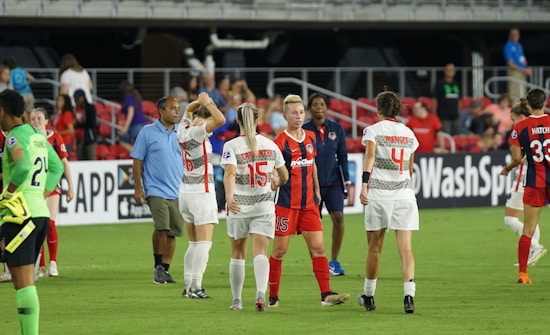 A group of female soccer players is standing on the field, some wearing white uniforms with red horizontal stripes and others wearing red and blue striped uniforms. The players appear to be interacting, with one player in white raising her hand near her face. Other individuals are visible in the background near the benches, some spectators sitting in the stands, and a coach or staff member in a blue shirt.