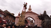 A large crowd gathers in front of a historical archway adorned with intricate carvings, witnessing a cultural or religious event. A tall straw structure decorated with fabric and ornaments stands prominently, with a mythical creature statue nearby. The atmosphere is festive, with people wearing traditional attire.