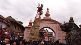 A large crowd gathers in front of a historical archway adorned with intricate carvings, witnessing a cultural or religious event. A tall straw structure decorated with fabric and ornaments stands prominently, with a mythical creature statue nearby. The atmosphere is festive, with people wearing traditional attire.