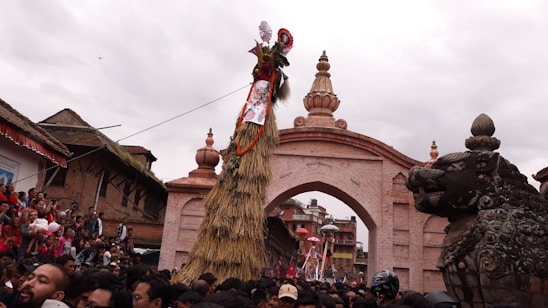 A large crowd gathers in front of a historical archway adorned with intricate carvings, witnessing a cultural or religious event. A tall straw structure decorated with fabric and ornaments stands prominently, with a mythical creature statue nearby. The atmosphere is festive, with people wearing traditional attire.