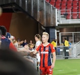 A soccer player wearing a red and blue striped jersey is interacting with fans, reaching over a barrier to shake hands. The player has short, styled blonde hair and the number 5 on her shorts. Behind her, a person in a white jersey is visible. The scene is set in a stadium with red seating and security personnel in the background.