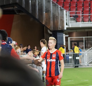 A soccer player wearing a red and blue striped jersey is interacting with fans, reaching over a barrier to shake hands. The player has short, styled blonde hair and the number 5 on her shorts. Behind her, a person in a white jersey is visible. The scene is set in a stadium with red seating and security personnel in the background.