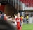 A soccer player wearing a red and blue striped jersey is interacting with fans, reaching over a barrier to shake hands. The player has short, styled blonde hair and the number 5 on her shorts. Behind her, a person in a white jersey is visible. The scene is set in a stadium with red seating and security personnel in the background.