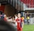 A soccer player wearing a red and blue striped jersey is interacting with fans, reaching over a barrier to shake hands. The player has short, styled blonde hair and the number 5 on her shorts. Behind her, a person in a white jersey is visible. The scene is set in a stadium with red seating and security personnel in the background.