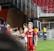 A soccer player wearing a red and blue striped jersey is interacting with fans, reaching over a barrier to shake hands. The player has short, styled blonde hair and the number 5 on her shorts. Behind her, a person in a white jersey is visible. The scene is set in a stadium with red seating and security personnel in the background.