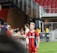A soccer player wearing a red and blue striped jersey is interacting with fans, reaching over a barrier to shake hands. The player has short, styled blonde hair and the number 5 on her shorts. Behind her, a person in a white jersey is visible. The scene is set in a stadium with red seating and security personnel in the background.