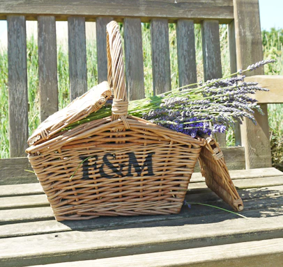 A wicker picnic basket with the letters 'F & M' is resting on a wooden bench. The basket contains bundles of fresh lavender flowers.