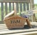 A wicker picnic basket with the letters 'F & M' is resting on a wooden bench. The basket contains bundles of fresh lavender flowers.