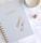 A spiral-bound notebook with a cover labeled NOTEWORTHY is placed on a marble surface. To its right are a pair of dangling gold earrings and a stack of rose gold rings. A candle sits near the top edge of the image.