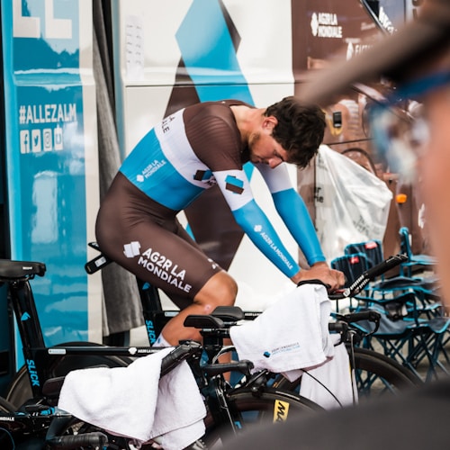 A cyclist wearing a brown and blue racing suit is bent over in a focused pose, positioned on a stationary bicycle surrounded by towels. The setting appears to be a team area, indicated by branding visible on the vehicle behind him. The scene conveys a sense of preparation and concentration common in competitive sports events.