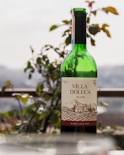 A green wine bottle with a label reading 'Villa Doluca Klasik' stands outdoors. The label features an illustration of a vineyard landscape. In the background, blurred greenery and a railing are visible, suggesting an outdoor setting.