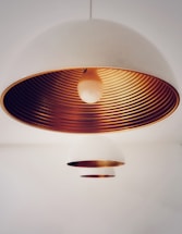 A close-up view of two dome-shaped pendant lights with a minimalist design. The larger lamp is prominently featured, showcasing its white exterior and striking metallic copper interior with concentric ring patterns. The smaller lamp is positioned further in the background, maintaining the same design aesthetic but appearing less detailed due to the focus on the main lamp.