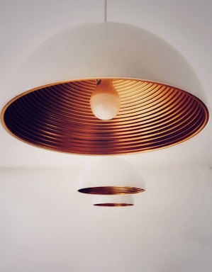 A close-up view of two dome-shaped pendant lights with a minimalist design. The larger lamp is prominently featured, showcasing its white exterior and striking metallic copper interior with concentric ring patterns. The smaller lamp is positioned further in the background, maintaining the same design aesthetic but appearing less detailed due to the focus on the main lamp.