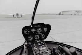 A helicopter cockpit with various instruments and gauges visible. The view includes a runway with an aircraft in the distance and a hangar building to the side.