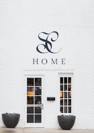 A minimalist white brick wall featuring a stylish monogram and the word 'HOME' above a modern glass-paneled door with two matching windows on each side. Large black planters filled with small plants flank the entrance, adding a touch of nature to the clean and elegant façade.