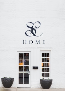 A minimalist white brick wall featuring a stylish monogram and the word 'HOME' above a modern glass-paneled door with two matching windows on each side. Large black planters filled with small plants flank the entrance, adding a touch of nature to the clean and elegant façade.