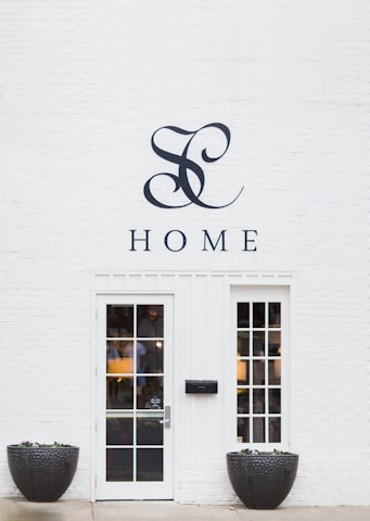 A minimalist white brick wall featuring a stylish monogram and the word 'HOME' above a modern glass-paneled door with two matching windows on each side. Large black planters filled with small plants flank the entrance, adding a touch of nature to the clean and elegant façade.