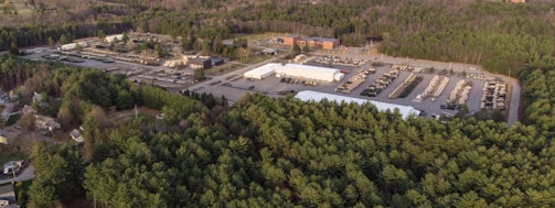 Aerial view of a large area with dense forest surrounding an industrial complex, which includes multiple buildings, storage units, and many parked vehicles. The scene shows a contrast between the green forested area and the structured, developed section with organized rows of similar vehicles.