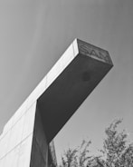 A black and white image of a modern architectural structure with a prominent overhanging concrete element. The word 'SAM' is engraved on the overhang. Sparse trees are visible below, and the sky is clear.
