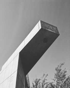 A black and white image of a modern architectural structure with a prominent overhanging concrete element. The word 'SAM' is engraved on the overhang. Sparse trees are visible below, and the sky is clear.
