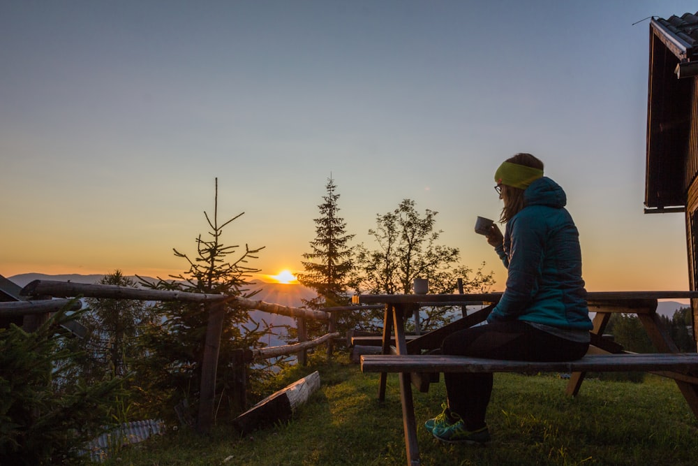 Frau in blauer Bubble-Jacke sitzt und trinkt einen Kaffee auf dem Berg während des Sonnenaufgangs