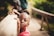 A young child wearing a pink shirt is looking directly at the camera while holding onto a wooden railing. The background features a blurred outdoor setting with greenery, suggesting a natural environment.
