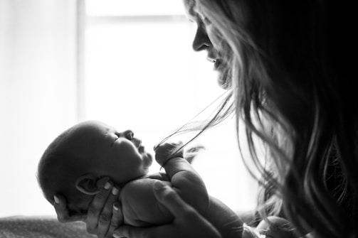 A black and white photograph of a woman holding a newborn baby close to her face. The baby is cradled in the woman's arms, and there is a sense of tenderness as she gazes lovingly at the child. The image captures the intimate bond between them, highlighted by the gentle play of light and shadow.
