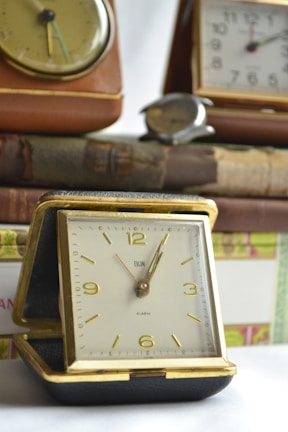 A collection of vintage clocks is displayed on a surface. The central focus is a gold-framed alarm clock with elegant numerals and hands, resting on a pile of old hardcover books. Other clocks, with classic designs, are arranged in the background, contributing to a nostalgic atmosphere. The lighting is soft, highlighting the textures and details of the objects.