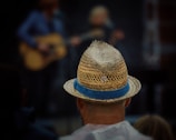 A person wearing a straw hat with a blue band stands in the foreground, facing away. In the blurred background, another person is holding a guitar, suggesting a musical performance.