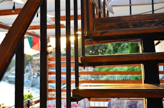 A wooden staircase with black metal railings is illuminated by warm sunlight. The background features a scenic view of a riverside or lakeside area with trees and a glimpse of a town or village. A section of a tricolor flag is visible, and there are small light bulbs hanging above.