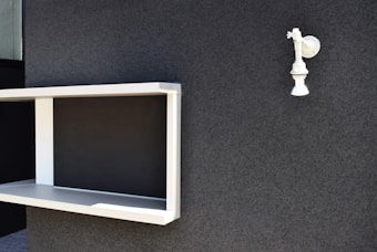 A minimalist setup with a white, wall-mounted shelf and a single spotlight fixture against a textured dark gray wall. The shelf is rectangular with clean lines and an open front. The spotlight fixture is positioned above and to the right of the shelf.