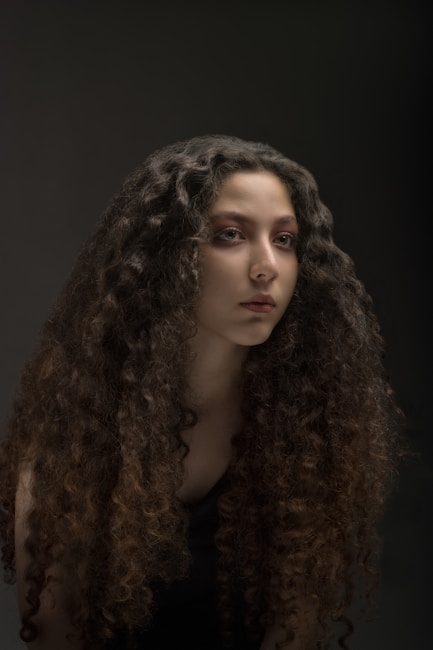 A person with long, curly hair gazes slightly away from the camera, set against a dark, neutral background. The lighting is soft and directed to highlight the textures and curls of the hair.